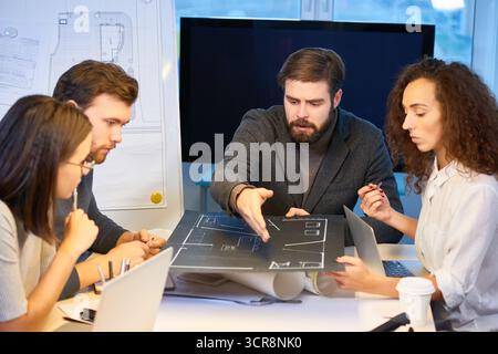 Architectes travaillant sur le projet de maison. Chef barbu en costume pointant le projet de la nouvelle maison faite par une jeune femme bouclée spécialiste et suggérant quelques changements Banque D'Images