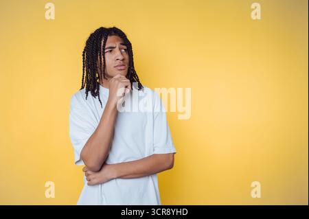 Réfléchi jeune homme afro-américain avec des cheveux tressés pose sur un fond jaune vif, réfléchissant sur les questions de la vie Banque D'Images