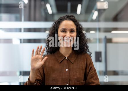 Jeune femme souriante portant un casque et agitant devant la caméra, participant à un appel vidéo ou fournissant un service client dans un environnement de bureau moderne Banque D'Images