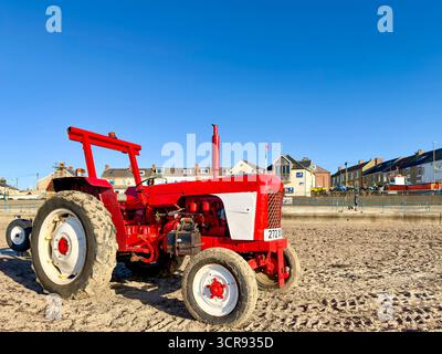 Tracteur rouge vintage David Brown garé sur une plage de sable avec des bâtiments de la ville côtière et un ciel bleu vif en arrière-plan Banque D'Images