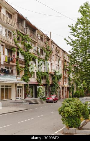 Vue d'une voiture rouge garée dans une rue bordée de bâtiments altérés drapés de vignes vertes vibrantes, créant un contraste saisissant, Goris, province de Syunik, Arménie. Banque D'Images