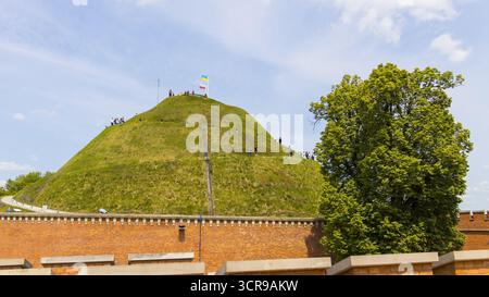 Vue d'un monticule vert vibrant se dresse haut derrière un mur de briques altérées, couronné d'un drapeau sous un ciel lumineux, Kraków, petite Pologne Voïvodie, po Banque D'Images