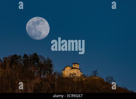 Vue aérienne d'une pleine lune lumineuse accroche haut au-dessus d'un bâtiment doré perché au sommet d'une colline, sa silhouette frappée contre le ciel assombrissant, Lugano, Suisse Banque D'Images