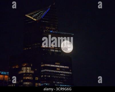 Vue aérienne d'une pleine lune rayonnante placée derrière le pinacle illuminé d'un gratte-ciel contre le ciel nocturne encreur, New York, New York, États-Unis. Banque D'Images