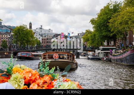 Amsterdan, pays-Bas - 5 août 2025 : des fleurs colorées au premier plan encadrent des vues panoramiques sur le canal et les bateaux à Amsterdam. Banque D'Images