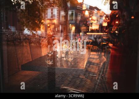 Table de restaurant vide avec des verres à vin et des arrangements floraux comme vu à travers la fenêtre en verre de l'extérieur. Lumière de fin de soirée, faible profondeur de f Banque D'Images