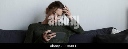 Une jeune femme est assise sur un canapé, profondément dans la pensée, vérifiant son téléphone avec une expression triste. Banque D'Images
