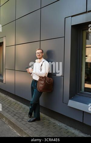 Un jeune enseignant se tient près d'un mur élégant, tenant un téléphone et exsudant un professionnalisme. Banque D'Images