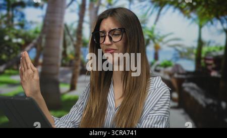 Femme tenant la tablette avec la main levée dans le parc vert parmi les palmiers portant une chemise rayée ; rejet dégoût aversion. Banque D'Images