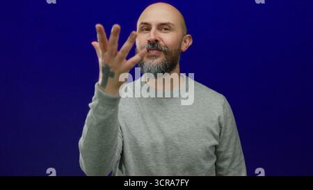 Des gestes d'homme chauve barbu sur fond bleu portant un pull gris exsudant charme et confiance. Banque D'Images