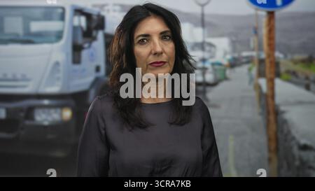 Femme hispanique d'âge moyen se tient debout montrant le visage dans une rue pluvieuse de la ville tout en portant un chemisier sombre ; sérieux reflet calme. Banque D'Images