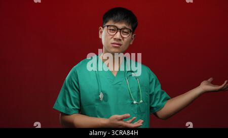Jeune homme dans des gommages verts sur un fond rouge vif gestes avec les deux mains comme si vous présentiez quelque chose, transmettant un professionnel médical curieux s Banque D'Images