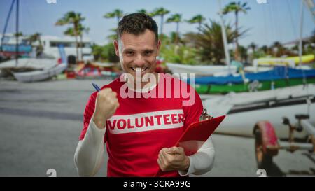 Homme portant une chemise rouge de volontaire debout sur le front de mer tenant une planche à pince montrant des pouces vers le haut expression confiante au milieu des palmiers et des bateaux suggérant outd Banque D'Images