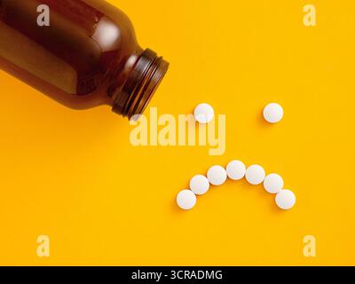 Visage triste fait de pilules débordant d'une bouteille de médicament représentant la dépression, les effets secondaires et la tristesse sur un fond jaune Banque D'Images