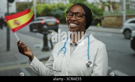 Femme habillée en médecin avec un stéthoscope souriant et tenant le drapeau espagnol dans une rue de la ville montrant la fierté nationale et l'identité professionnelle dans une urba Banque D'Images
