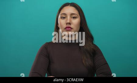 Jeune femme hispanique portant un haut noir avec les bras croisés et regardant latéralement dans le studio avec fond sarcelle ; rejet. Banque D'Images