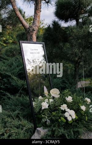 Miroir encadré foncé utilisé comme élégant signe de bienvenue de mariage, placé dans le jardin et décoré avec des arrangements floraux blancs et verts. Traduction de Ukr Banque D'Images