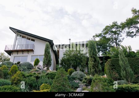 Maison blanche contemporaine avec de grandes fenêtres et un balcon, entourée d'un magnifique paysage en couches avec des conifères, des arbustes et des pierres Banque D'Images