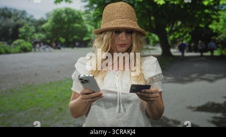 Femme tenant smartphone et carte de crédit tout en vérifiant une transaction dans la rue dans un parc vert portant chapeau de paille et chemisier blanc ; concentration. Banque D'Images