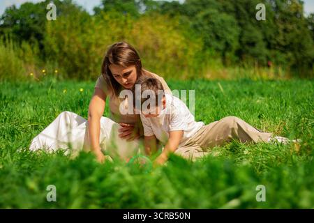 Mère aidant son petit fils à lire un livre sur les voitures et les transformateurs passant du temps sur la nature assis sur l'herbe le jour d'été. Banque D'Images
