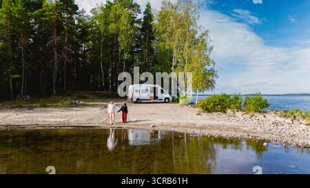 Deux amis se promènent le long de la rive sablonneuse du lac Saimaa, entouré d'une végétation luxuriante et d'une atmosphère tranquille. Un camping-car est garé à proximité, prêt pour l'exploration. Banque D'Images