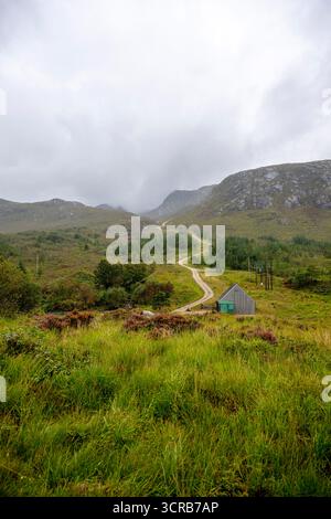 Un paysage rural tranquille avec un chemin de terre sinueux menant à une petite cabane en bois, situé au milieu de collines verdoyantes et d'arbres dispersés sous un lourd overca Banque D'Images