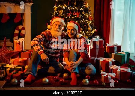 Un joyeux couple retraité dans des pulls assortis est assis avec des cadeaux à côté d'un arbre de noël brillant entouré de cadeaux et d'un éclairage chaleureux créant un confortable Banque D'Images