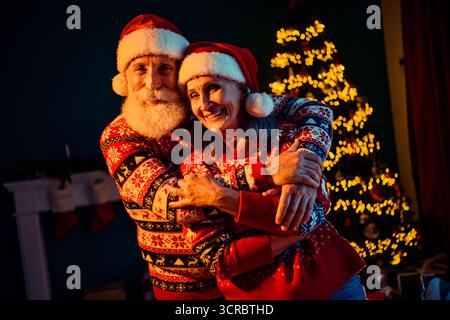 Couple en chandails de Noël portant des chapeaux de Père Noël câlin dans une maison confortable à côté d'un arbre de Noël scintillant avec des lumières festives créant des vacances chaudes Banque D'Images