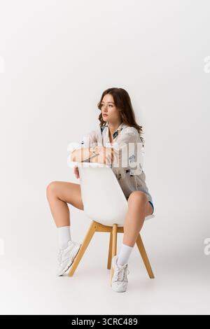 Jeune femme à la mode prend une pose confiante avec une chaise, mettant en valeur son style unique. Banque D'Images