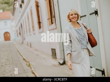 Une femme confiante marche le long d'une étroite rue pavée entourée de beaux bâtiments historiques. Elle porte un blazer léger et une chemise rayée, enjo Banque D'Images