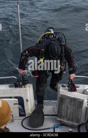 Mer de Barents, Russie - 14 juin 2025 : un plongeur en combinaison sèche noire avec équipement de plongée remonte sur un bateau après une activité sous-marine Banque D'Images