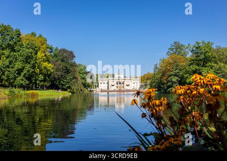 Le palais sur l'île (polonais : Palac Na Wyspie) dans le parc des bains royaux de Varsovie, vue paysage avec le lac, arbres et fleurs d'automne orangées, ciel bleu Banque D'Images