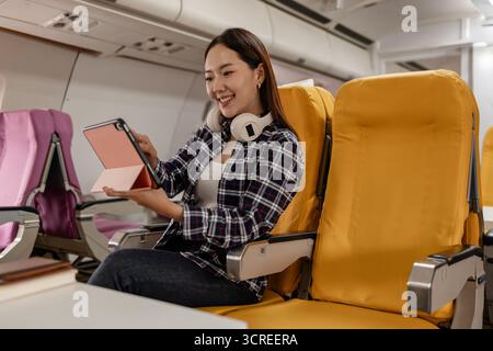 Jeune femme asiatique assise dans un siège d'avion avec couverture jaune, souriant tout en utilisant la tablette pendant le vol. Elle porte une tenue décontractée avec des écouteurs autour Banque D'Images