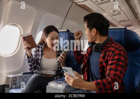 Jeune couple asiatique excité assis dans un avion, tenant un passeport, de l'argent et un smartphone tout en célébrant ensemble. La femme a l'air heureuse, et le Banque D'Images