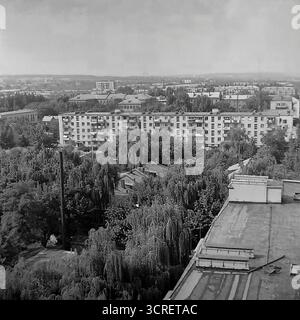 Une photographie panoramique panoramique en noir et blanc des années 1980, montrant le paysage urbain de Sloviansk, en RSS d'Ukraine, depuis un point de vue élevé. La vue capture la zone autour de Svobody Street, présentant une tapisserie urbaine diversifiée caractéristique de l'ère soviétique. Le paysage est un mélange d'immeubles à panneaux de plusieurs étages, de bâtiments administratifs plus anciens et d'un vaste quartier résidentiel de faible hauteur avec des maisons privées nichées au milieu d'une végétation luxuriante. Au loin, un horizon industriel avec des cheminées d'usine enracine fermement la ville dans le cœur industriel du Donbass Banque D'Images