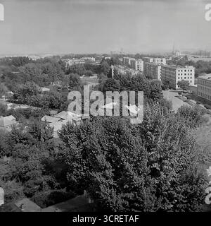 Une photographie panoramique panoramique en noir et blanc des années 1980, montrant le paysage urbain de Sloviansk, en RSS d'Ukraine, depuis un point de vue élevé. La vue capture la zone autour de Svobody Street, présentant une tapisserie urbaine diversifiée caractéristique de l'ère soviétique. Le paysage est un mélange d'immeubles à panneaux de plusieurs étages, de bâtiments administratifs plus anciens et d'un vaste quartier résidentiel de faible hauteur avec des maisons privées nichées au milieu d'une végétation luxuriante. Au loin, un horizon industriel avec des cheminées d'usine enracine fermement la ville dans le cœur industriel du Donbass Banque D'Images