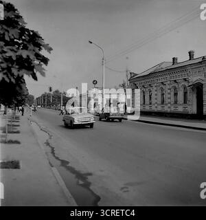 Une scène de rue d'archives en noir et blanc des années 1980 sur la rue Iskry (aujourd'hui rue Shovkovychna) à Sloviansk, RSS d'Ukraine. La photographie capture un mélange unique d'époques, avec des voitures classiques de l'ère soviétique, y compris une Moskvitch et une VAZ Zhiguli, passant devant une rangée de maisons de marchands du XIXe siècle bien conservées. Les façades en briques ornées des bâtiments de l'époque tsariste contrastent avec les véhicules et les infrastructures de l'époque socialiste. Cette image est un regard nostalgique sur la vie quotidienne dans une ville soviétique provinciale, où différentes couches historiques coexistaient. Banque D'Images