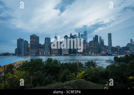 Vue de l'horizon de la ville enflammée de lumière, reflétant dans les eaux ci-dessous, encadrée par des arbres verdoyants sous un vaste ciel, strié de nuages, New York, New York, États-Unis. Banque D'Images