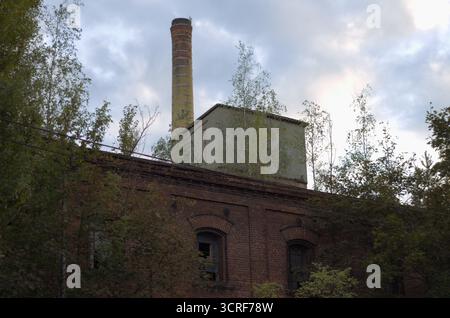 Ancien mur de briques d'usine avec fenêtres cintrées, arbres. Smokestack en arrière-plan. Ciel ensoleillé avec des nuages, Pologne Banque D'Images
