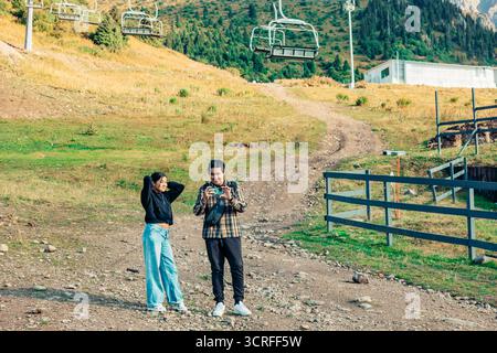 Une jeune femme asiatique aux longs cheveux noirs et un jeune homme hispanique aux courts cheveux noirs se tiennent sur un chemin de montagne près d'un téléphérique. Ils s'amusent Banque D'Images