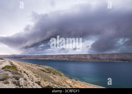 Vue sur les eaux sombres de la mer Adriatique rencontrant la côte accidentée sous un ciel spectaculaire rempli de nuages tourbillonnants, Pag, comté de Zadar, Croatie. Banque D'Images