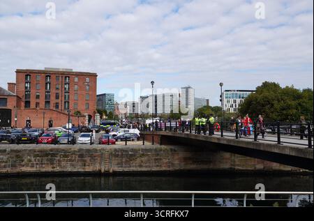 Vue sur Albert Dock à Liverpool avec des entrepôts historiques en briques rouges, des voitures garées, des bâtiments modernes et des gens marchant sur un pont par une journée nuageuse Banque D'Images