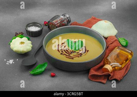 Soupe crémeuse à la courge pattypan, garnie de basilic et de glaçage balsamique, servie dans un bol tendance avec des ingrédients frais et des épices. Béton de pierre sombre b Banque D'Images