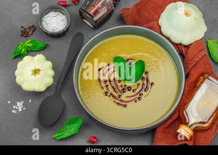 Soupe crémeuse à la courge pattypan, garnie de basilic et de glaçage balsamique, servie dans un bol tendance avec des ingrédients frais et des épices. Béton de pierre sombre b Banque D'Images