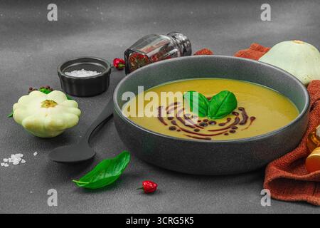 Soupe crémeuse à la courge pattypan, garnie de basilic et de glaçage balsamique, servie dans un bol tendance avec des ingrédients frais et des épices. Béton de pierre sombre b Banque D'Images