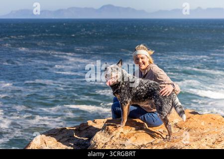 Femme assise sur un rocher avec un chien de bétail australien (Blue Heeler) regardant la caméra et souriant, explorant en plein air avec l'océan et la moun Banque D'Images