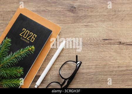 Un espace de travail bien rangé avec un planificateur 2026, un cahier, un crayon blanc, des verres sur un bureau en bois, suggérant planification, concentration et matinée productive. Banque D'Images