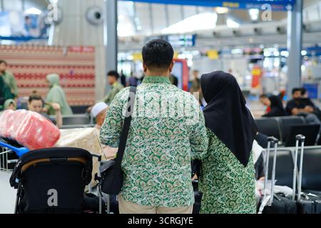 Un couple vêtu de chemises à motifs verts assortis se tient debout avec leurs bagages à l'aéroport. Les membres de leur famille et d'autres voyageurs sont visibles dans le Banque D'Images