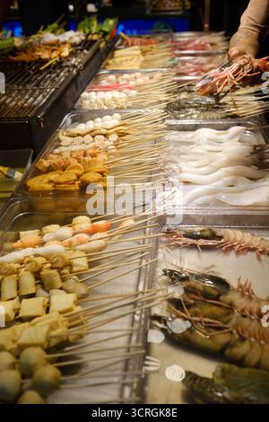 Un étal de marché est rempli d'un assortiment de brochettes de fruits de mer et de viandes. Un vendeur prépare des brochettes fraîches dans une atmosphère animée pendant un même Banque D'Images