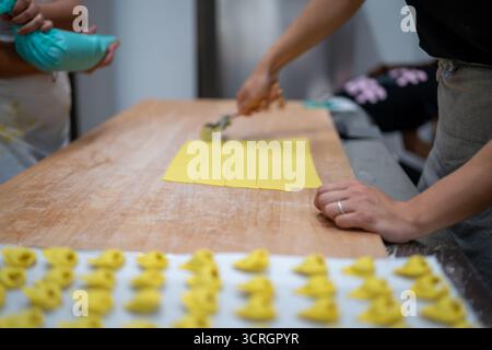 Deux personnes préparent soigneusement des pâtes fraîches faites maison sur une grande table en bois avec un rouleau à pâtisserie créant des carrés parfaits Banque D'Images